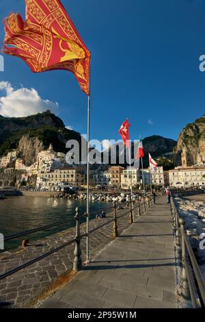 Drapeau vénitien en volant sur un mât entre d'autres drapeaux sur la jetée principale à Amalfi, Salerno, Italie avec la ville derrière. Côte amalfitaine. Mer Tyrrhénienne. Les gens sur la jetée. Banque D'Images