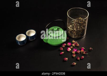 Une petite théière en fonte verte de type asiatique avec du thé sec dans un bol en verre, des tasses en porcelaine et des boutons-pression Banque D'Images