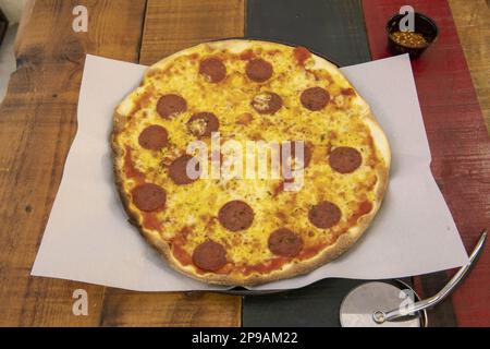 Une pizza à croûte mince fraîchement cuite avec beaucoup de fromage, de pepperoni et un couteau à pizza en acier inoxydable Banque D'Images