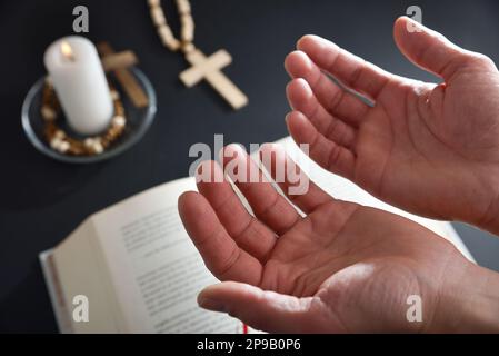 Christian priant avec les mains vers le haut avec la table noire dans le fond avec la bible croix chrétienne et la bougie brûlante. Banque D'Images