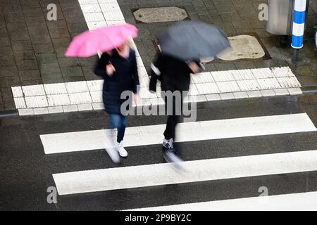 flou de mouvement de deux piétons avec des parapluies noirs et roses traversant un passage de côté sous la pluie Banque D'Images