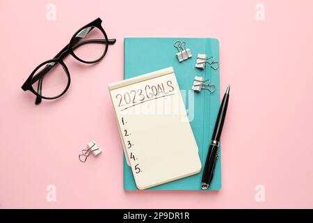 Carnet avec inscription 2023 objectifs, stylo et lunettes sur fond rose, plat. Objectifs de la nouvelle année Banque D'Images