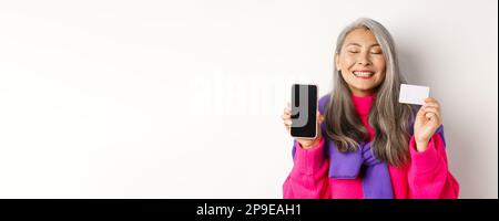 Achats en ligne. Heureuse et ravie asiatique femme senior souriant avec joie, montrant l'écran vide de smartphone et carte en plastique, l'achat dans Internet, blanc Banque D'Images