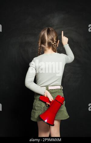 Jolie petite fille tenant un mégaphone et pointant vers le haut contre un tableau noir. Enfant avec haut-parleur Banque D'Images