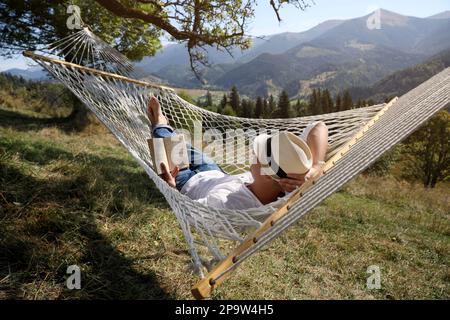 Un jeune homme lisant un livre dans un hamac en plein air par beau temps Banque D'Images