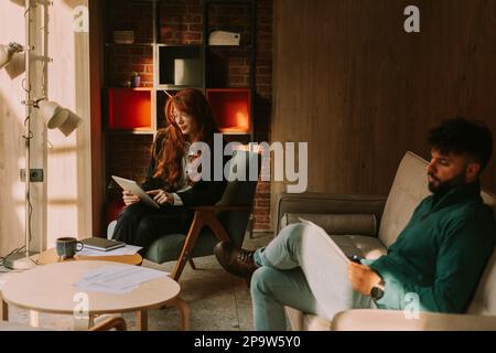 Un homme d'affaires élégant et élégant qui lit le rapport financier quotidien pendant que sa collègue Ginger travaille sur sa tablette Banque D'Images