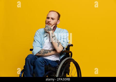 Un homme en fauteuil roulant a des problèmes avec le système musculo-squelettique regarde la caméra avec soin avec des tatouages sur ses bras assis sur un studio jaune Banque D'Images