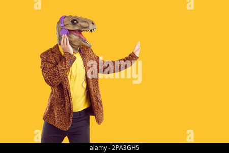 Doudoudoudoudoueur excentrique en caoutchouc masque de dinosaure écouter de la musique dans les écouteurs sur fond jaune. Banque D'Images