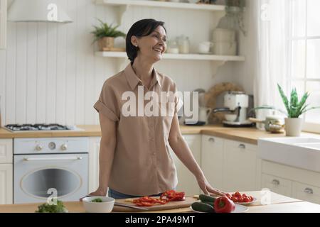 Une femme de chef de 50s ans gaie prépare une salade à la table de la cuisine Banque D'Images