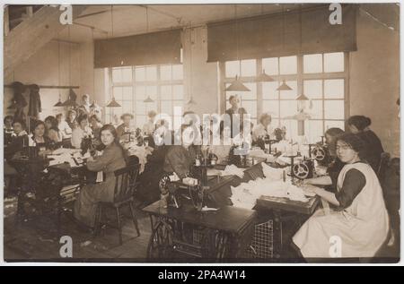 Photo du début du 20th siècle d'un groupe de travailleurs du textile. Les femmes sont assises aux machines à coudre à pédales dans un atelier avec une grande fenêtre Banque D'Images