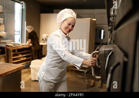 Femme souriante boulangère ouvrant un grand four industriel dans la cuisine moderne de boulangerie Banque D'Images