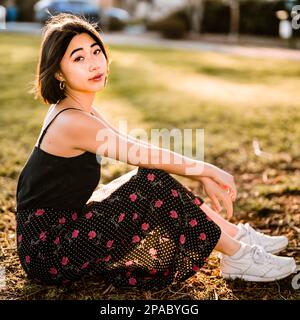 Belle jeune femme asiatique assise sur l'herbe dans un parc au coucher du soleil | vue latérale | appareil photo en face | rétroéclairage Banque D'Images