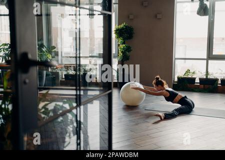 Sportive flexible femme avec les cheveux dans un petit pain étirant les jambes tout en étant assis en position séparée sur le tapis de yoga dans le studio de fitness, en utilisant le fitball et se détendre à l'arrière Banque D'Images