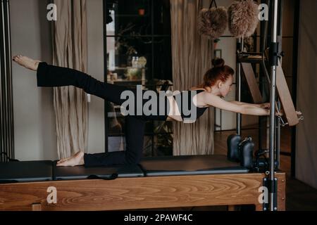 Vue latérale d'une jeune femme athlétique qui s'étire les jambes sur une machine à pilates ou un reformeur Cadillac, en portant un haut et un pantalon noirs tout en faisant de l'exercice en salle de sport ou en salle de sport Banque D'Images