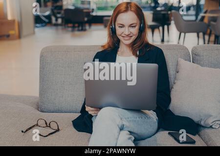 REDHEAD jeune femme communiquant avec les clients à l'aide d'un ordinateur portable et d'un casque sans fil, femme souriante parlant pendant un appel virtuel assis sur un canapé dans un café Banque D'Images