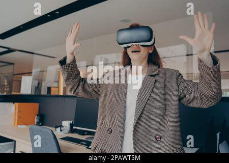 Femme avec une expression faciale surprise, qui vit un monde incroyable de réalité virtuelle avec des lunettes VR, debout seul devant le bureau, tenant Banque D'Images