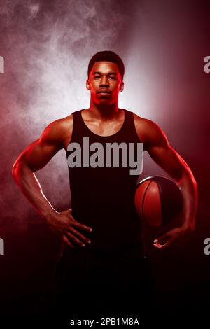 Côté joueur de basket-ball éclairé de couleur rouge tenant une balle sur fond de fumée. Sérieux homme afro-américain concentré. Banque D'Images