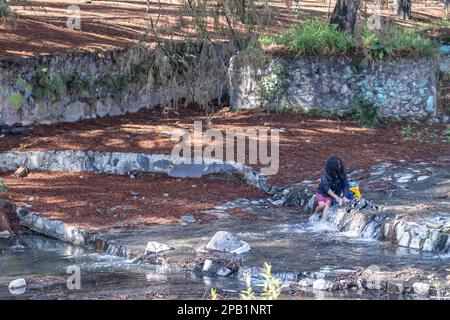 Zapopan, Jalisco Mexique. 1 janvier 2023. Ruisseau avec peu d'eau entre les murs de pierre, sans-abri personne assis sur des pierres laver ses vêtements, coulant Banque D'Images