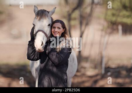 Jeune femme debout à côté de cheval blanc arabe souriant, le soleil brille sur les arbres flous et le fond du champ Banque D'Images