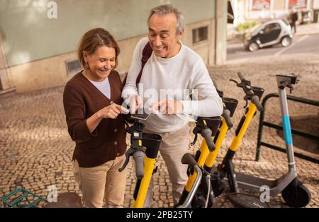 Heureux couple d'âge mûr en utilisant un smartphone louant des scooters électriques à l'extérieur Banque D'Images