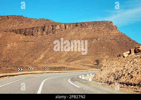 Courbe sur route asphaltée, petites montagnes de l'Atlas et buissons bas des deux côtés, ciel clair au-dessus - paysage typique dans le sud du Maroc Banque D'Images