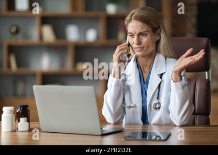 Femme stressée médecin parlant sur téléphone portable et utilisant l'ordinateur portable en clinique Banque D'Images