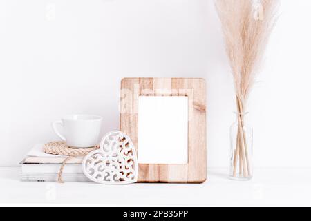 Cadre photo en bois maquette, livres, tasse à café, décor sculpté coeur et pampas herbe dans vase sur table blanche. Copier l'espace. Banque D'Images