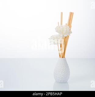 Photo dans le style du minimalisme. Vase blanc avec fleurs et bâtonnets de bambou sur fond miroir clair. Banque D'Images