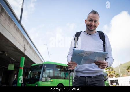 Un voyageur souriant tenant une carte et portant un sac à dos à une gare routière Banque D'Images