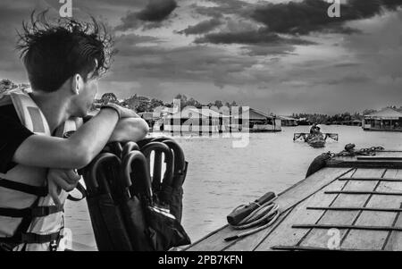 Un jeune homme sur un bateau en bois regarde un bateau de pêche traditionnel à côté de fermes de poissons sur le Mékong près de Tan Chau Vietnam. Banque D'Images