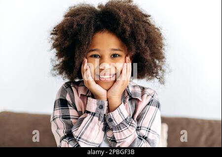 Photo d'une jolie petite fille afro-américaine d'âge préscolaire, avec des cheveux bouclés, dans un vêtement de base, assis sur le canapé, regarde l'appareil photo, sourit amicaux. Bonne enfance Banque D'Images