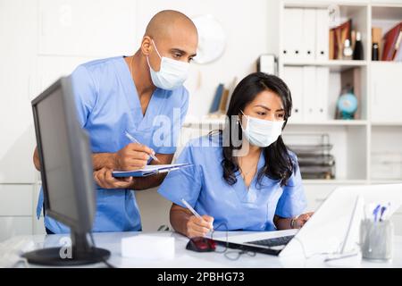 Twp médecins en masques chirurgicaux remplissant les formes médicales dans ordinateur portable Banque D'Images