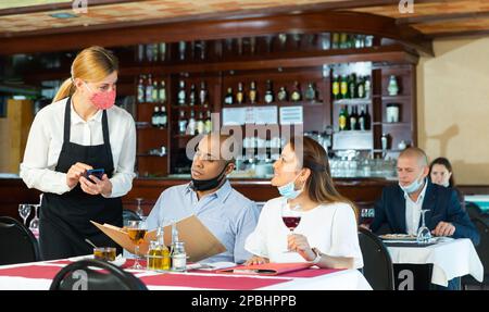 Une serveuse sympathique dans un masque de protection prend l'ordre des visiteurs du restaurant Banque D'Images