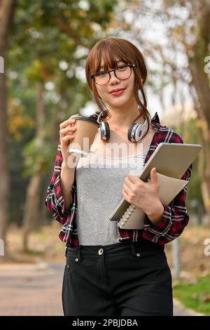 Portrait, souriant et jolie jeune asiatique étudiante à l'université dans des vêtements et des lunettes décontractés, se tient dans le parc du campus tenant sa tasse à café à emporter, Banque D'Images