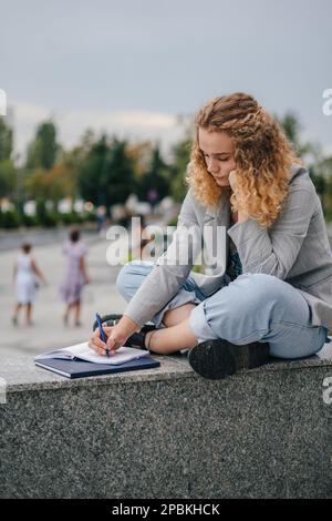 Jeune beau adolescent étudiant en plein air dans le parc écrivant dans le carnet , avec un regard sérieux concentré, faisant des devoirs. Prise de notes sur Banque D'Images