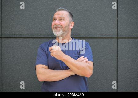 Homme d'âge moyen barbu regardant avec un sourire heureux posant à l'extérieur contre un mur Banque D'Images