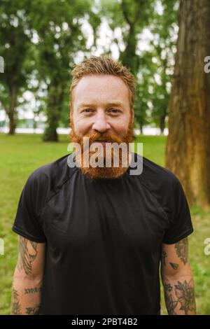Portrait de jeune beau barbu tatoué tête rouge souriant homme regardant l'appareil photo, tout en se tenant dans un parc vert Banque D'Images
