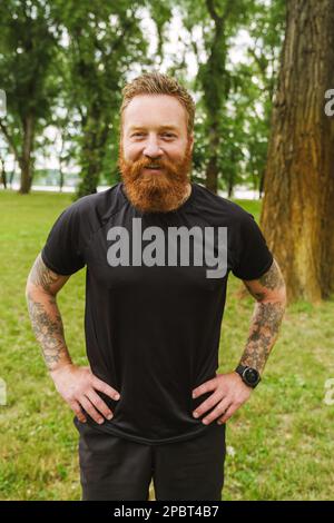 Portrait de jeune beau barbu tatoué tête rouge souriant homme regardant l'appareil photo, tout en se tenant dans un parc vert Banque D'Images