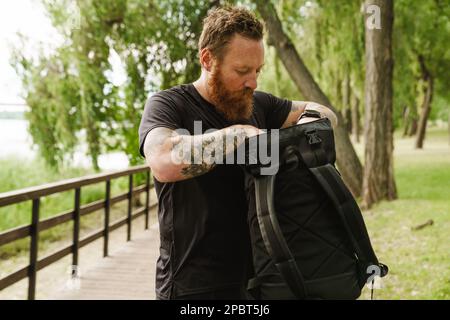 Jeune beau calme tatoué homme barbu rummaing dans son sac à dos en se tenant dans un parc ensoleillé Banque D'Images