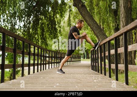 Jeune beau homme à barbe tatoué qui étire sa jambe sur une main courante en bois dans un parc ensoleillé Banque D'Images