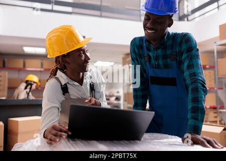 Souriant chefs de service de la logistique analysant le rapport de distribution du fret sur ordinateur portable. Les employés de l'entrepôt vérifient l'approvisionnement en stock sur ordinateur, rient et travaillent ensemble dans l'entrepôt Banque D'Images