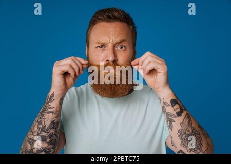 Portrait de l'adulte barbu tatoué beau homme élégant sérieux tenant sa grosse moustache et regardant l'appareil photo, tout en se tenant sur le dos bleu isolé Banque D'Images