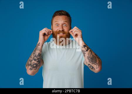 Portrait de l'adulte barbu tatoué beau homme stylé et enthousiaste tenant sa grosse moustache et regardant l'appareil photo, tout en se tenant sur le bleu isolé Banque D'Images