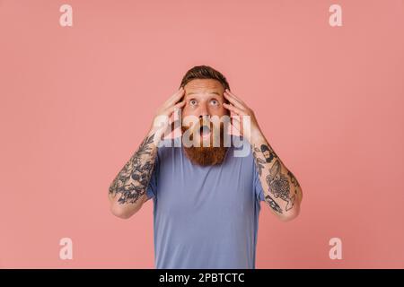 Portrait de l'adulte tête rouge barbu tatoué homme choqué dans le t-shirt avec la bouche ouverte touchant sa tête et regardant vers le haut , tout en se tenant au-dessus de l'isolat Banque D'Images