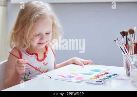 Une fille maussée dessine un dessin d'enfant avec des peintures et un pinceau. Banque D'Images