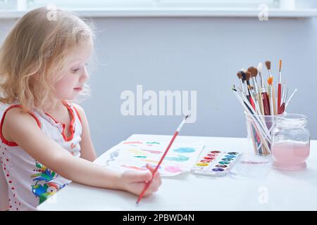 Une jolie fille aux cheveux bouclés peint à une petite table de jardin d'enfants. Banque D'Images