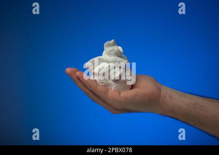 Mousse de rasage blanche maintenue dans la paume par un homme caucasien. Gros plan en studio, isolé sur fond bleu. Banque D'Images