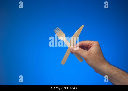 Couteau et fourchette en bois à usage unique, à usage unique et biodégradable, tenu en main par un homme caucasien. Gros plan en studio, isolé sur fond bleu. Banque D'Images