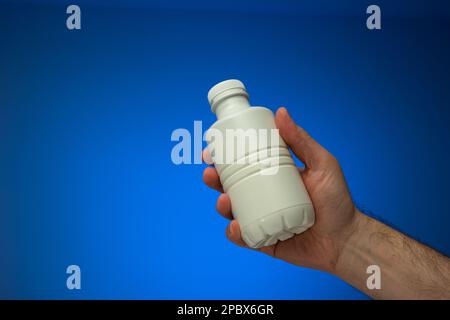 Flacon en plastique blanc opaque tenu à la main par un mâle caucasien. Gros plan en studio, isolé sur fond bleu. Banque D'Images