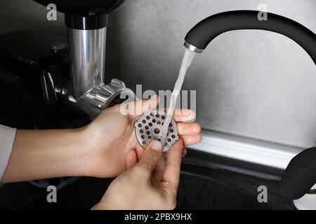 Femme lavant la plaque de coupe du moulin à viande électrique sous l'eau du robinet dans l'évier de cuisine à l'intérieur, gros plan Banque D'Images